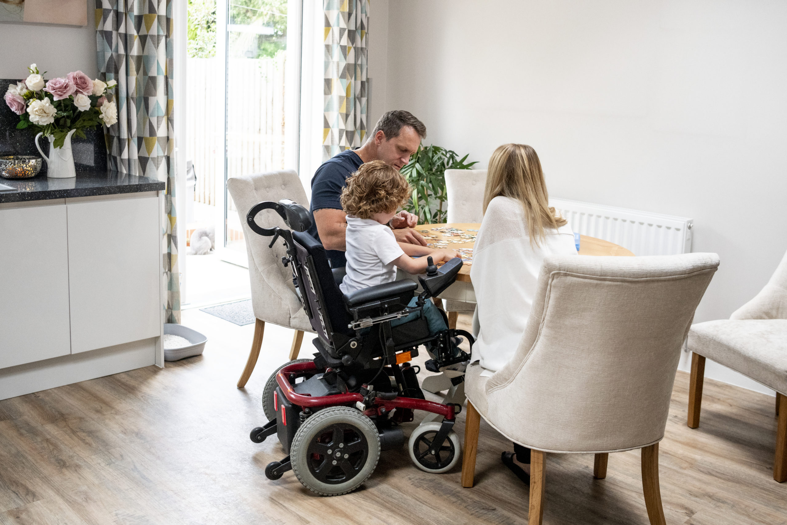 Kleinfamilie mit Kind im Elektro-Rollstuhl bei einem Spiel am Wohnzimmertisch