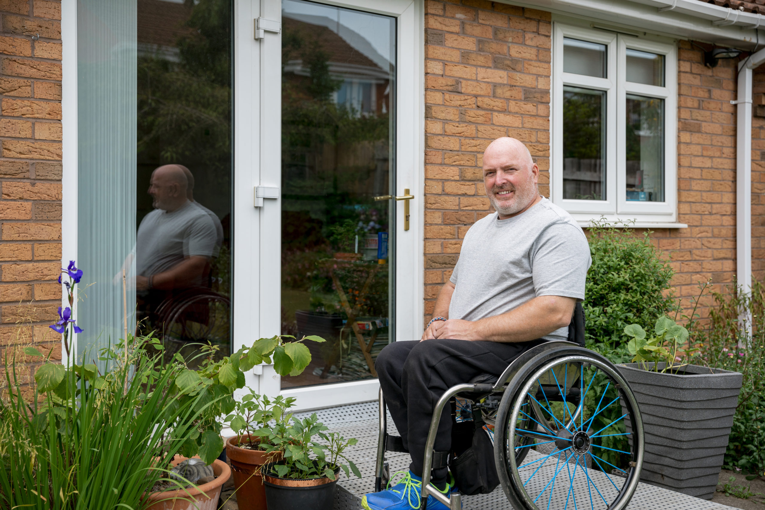 Lächelnder Mann im Rollstuhl auf der Terrasse vor Pflanzen und der Terrassentür